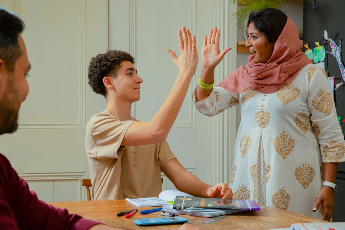 A foster carer and a child are sharing a high-five in a kitchen setting. The adult, a woman stands beside the seated child. The child, a teenage boy with curly hair, has an open textbook and a notebook on the table in front of him. Another person, partially visible on the left, is also seated at the table.