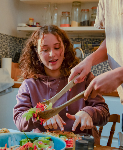 A young teenage girl sits at a kitchen table preparing her food, while another person stands out of shot but is using salad servers to lift some brightly coloured salad