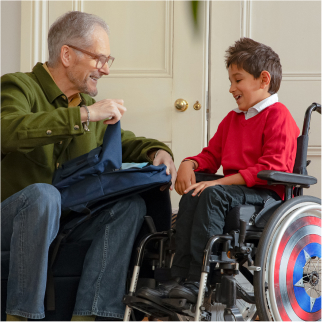 A young boy sits in a wheelchair smiling with an older male adult who is sat on the sofa. The adult is holding a rucksack open to look at the contents.