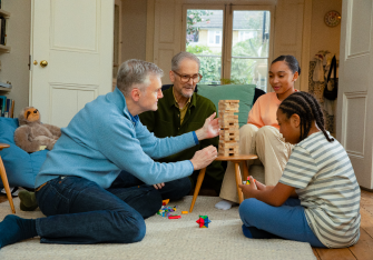 Two male adults and two children are sat on the floor playing a game of Jenga. They are all focused on one of the adults taking their turn at Jenga.