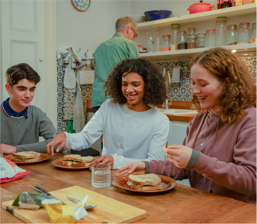 Three smiling teenagers sat around a kitchen table with sandwiches on the table in front on them, there is an adult in the background with his back to the camera who looks like they are preparing food.