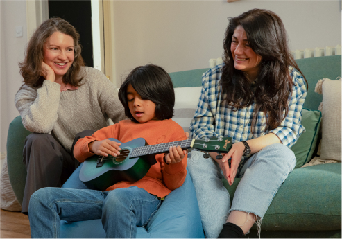 A young boy on a beanbag concentrating on playing a small blue guitar, sat in-between two smiling adult women who are sitting on a green sofa.