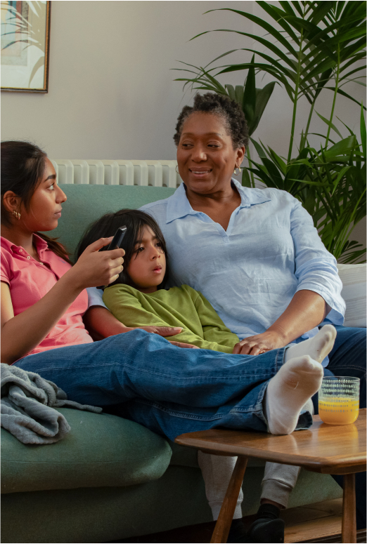 Woman sat on the sofa with 2 children, cuddling the younger child while looking with a smile at the older girl, who is sat holding a TV remote and has her legs resting on a table