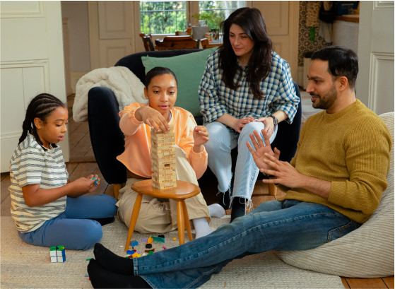 A family is gathered in a cozy living room, engaging in playful activities together. A young child is sitting on the floor, working on a Rubik's cube. The second child is kneeling and carefully placing a block on top of a Jenga tower. An adult woman seated on a couch, is watching the Jenga game with a smile. Another adult, a man, is sitting on the floor with his legs stretched out, clapping his hands in encouragement.