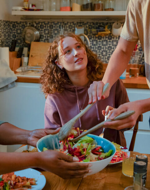 A teenage girl with curly hair is sitting at a wooden kitchen table, looking up to an adult who is holding salad servers and serving a colourful salad from a large bowl. There is another person out of the picture holding the bowl steady.