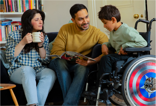 A young boy sits in a wheelchair with two adults who are sitting on the sofa. The male adult is holding a book for the boy to look at, and the female adult is smiling at the boy while holding a mug.