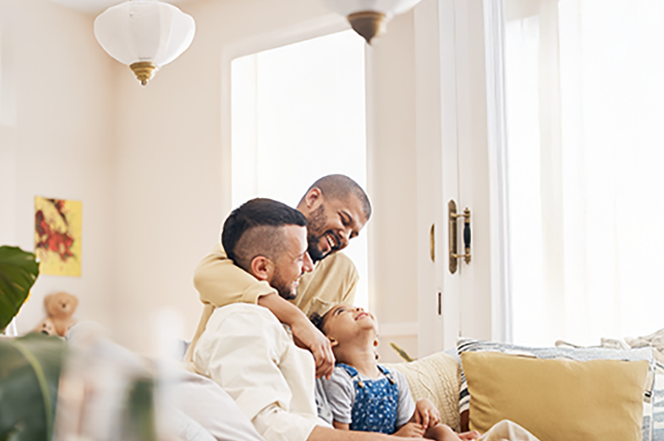 two men in a same sex couple hugging on the sofa with their young foster child