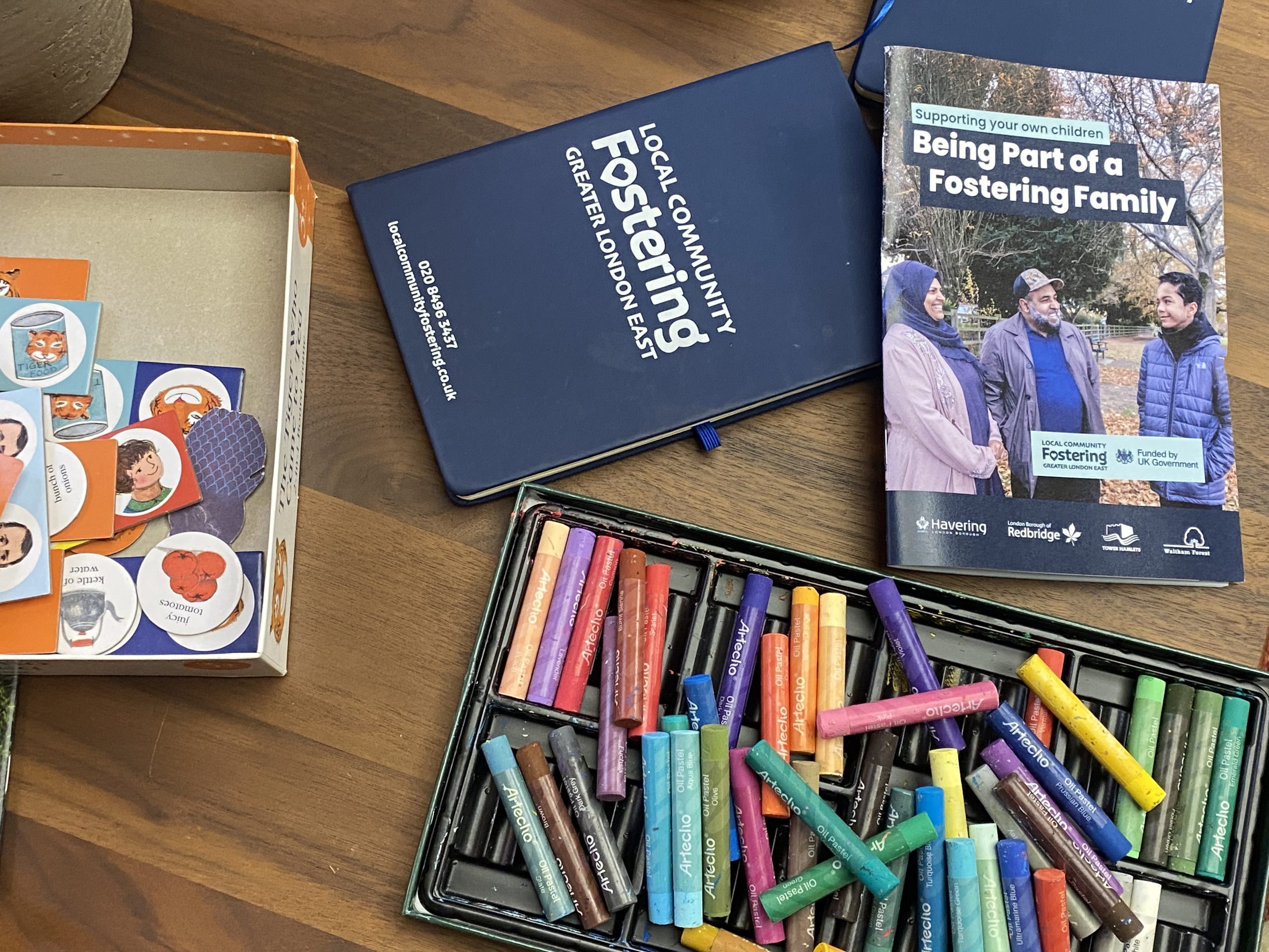 view of a table with coloured chalks, a board game, the Local Community Fostering notebook and one of the Being Part of a Fostering Family booklet.