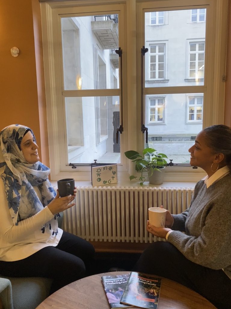two women of seemingly different faiths having a conversation over tea. A drawing of different faith symbols in the background.