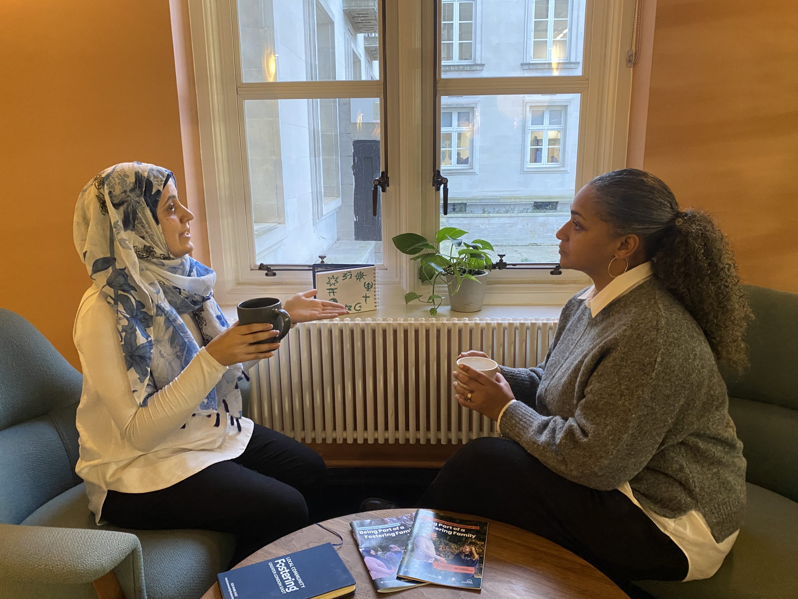 two women of seemingly different faiths having a conversation over tea. A drawing of different faith symbols in the background. This image is landscape.